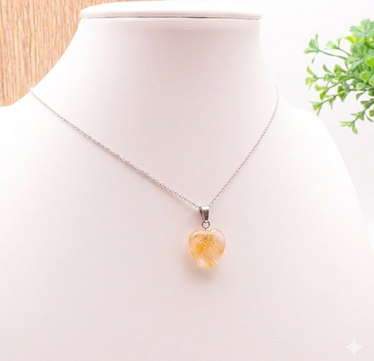 Un pendentif en forme de cœur en pierre naturelle jaune translucide, suspendu à une fine chaîne argentée sur un buste d'exposition blanc. Le décor est lumineux, avec un arrière-plan boisé à gauche et une touche de verdure discrète sur la droite. L'image met l'accent sur la clarté et la douceur des reflets de la pierre.