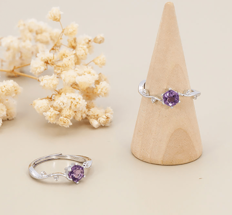 Une bague en argent ornée d'une améthyste ronde violette. Serti et pierre centrale : La pierre centrale est une améthyste facettée d'un violet vibrant, taillée en rond et maintenue par six griffes métalliques. Anneau et détails : L'anneau fin présente des ondulations d'inspiration végétale. Il est serti de quatre petits strass scintillants, répartis de part et d'autre de la pierre centrale le long de la tige. L'anneau est ajustable par le dessous. Mise en scène : Deux bagues identiques sont présentées sur un fond beige très clair. L'une est enfilée sur un cône de présentation en bois lisse, l'autre repose à plat au premier plan, à côté de petites fleurs séchées écrues (gypsophile).