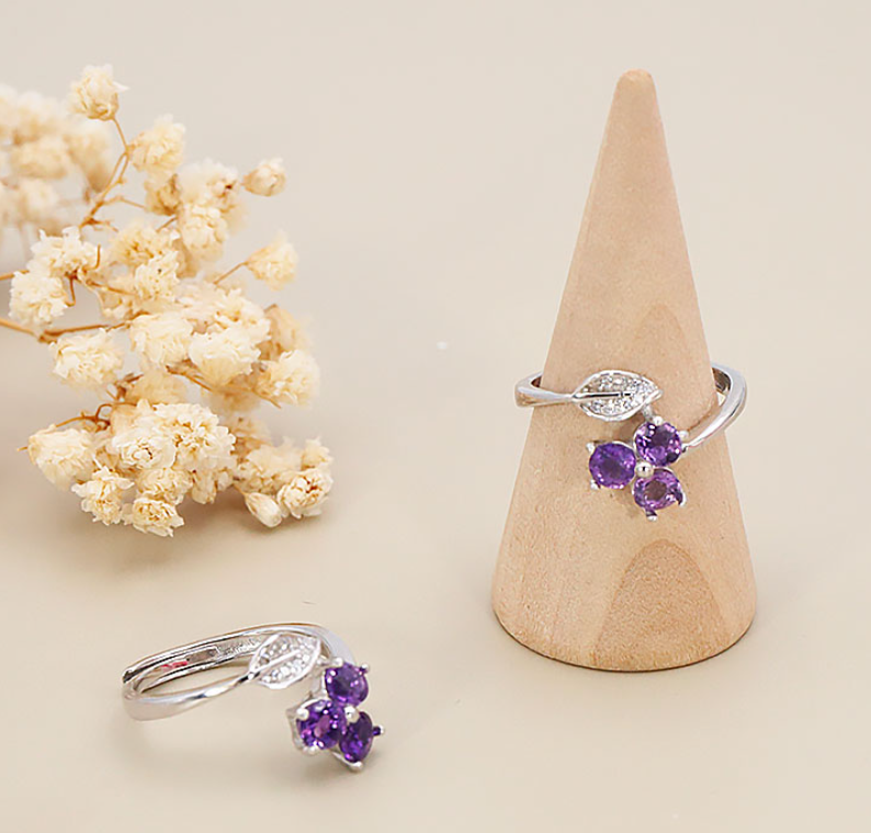 Photo de produit : Une bague réglable en argent ornée d'un motif floral délicat composé de trois améthystes violettes facettées et d'une petite feuille sertie de strass. Mise en scène : Le bijou principal est présenté verticalement sur un cône de support en bois clair, tandis qu'un second exemplaire est posé au premier plan à gauche. Arrière-plan : Le décor minimaliste et épuré est complété sur la gauche par une délicate branche de petites fleurs séchées de couleur blanche et crème.