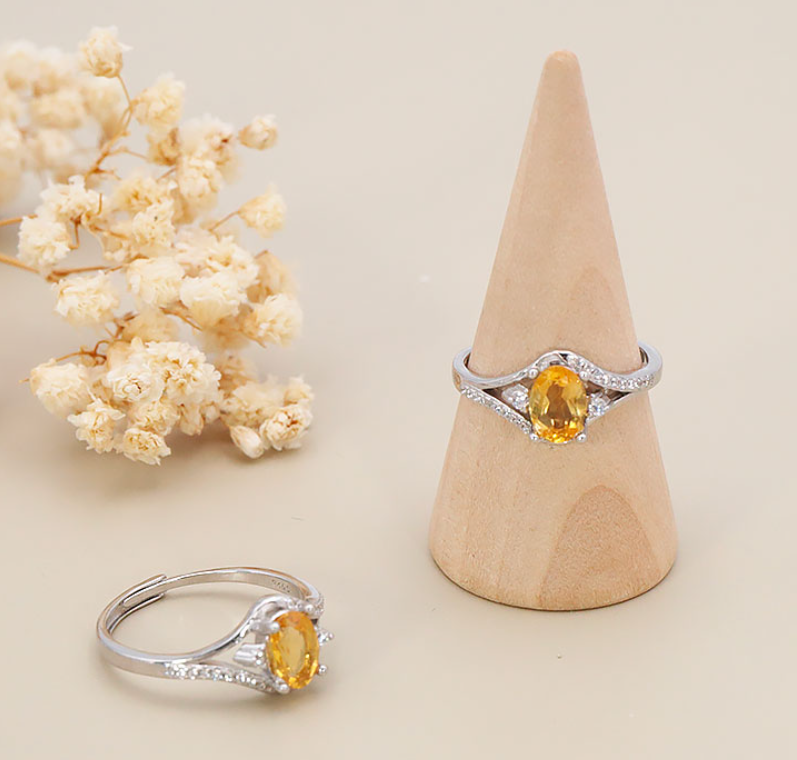 Photo de produit : Une bague réglable en argent ornée de citrine jaune facettées Mise en scène : Le bijou principal est présenté verticalement sur un cône de support en bois clair, tandis qu'un second exemplaire est posé au premier plan à gauche. Arrière-plan : Le décor minimaliste et épuré est complété sur la gauche par une délicate branche de petites fleurs séchées de couleur blanche et crème.