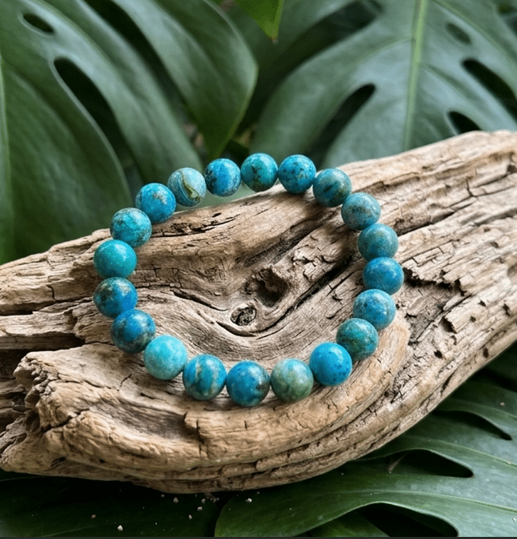 Un bracelet de perles rondes en turquoise, aux nuances bleues et vertes marbrées de brun, repose sur des morceaux de bois flotté gris et texturés. Deux grandes feuilles vert foncé de Monstera, perforées naturellement, encadrent le bijou sous une lumière naturelle.