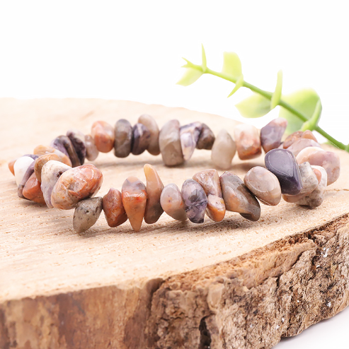 Ce bracelet circulaire est composé de petites pierres naturelles et polies de Tiffany Stone, qui présentent des formes irrégulières, lisses et galbées. Les perles dévoilent une mosaïque de couleurs douces et marbrées, mélangeant des nuances de violet, de mauve, de beige, de gris clair et de rose saumon. Le bijou est mis en valeur à plat sur un support en bois brut à l'écorce visible, le tout se détachant sur un fond blanc épuré où l'on distingue une petite pousse végétale verte et floutée.