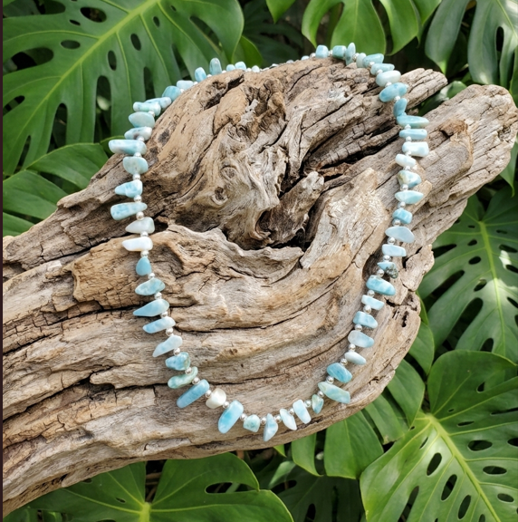 Ce collier en Larimar est composé de petites pierres naturelles roulées aux nuances bleu ciel et marbrées de blanc. Il est harmonieusement disposé en cercle par-dessus une grosse branche de bois flotté texturée et claire. L'ensemble repose devant un arrière-plan végétal dense, formé par de grandes feuilles vertes et découpées de Monstera.