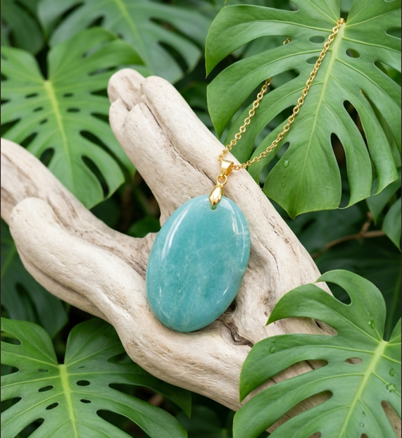 Un pendentif en pierre bleu turquoise, lisse et ovale, est suspendu à une fine chaîne dorée et posé sur un morceau de bois clair aux formes naturelles. Autour, de grandes feuilles vertes tropicales aux découpes profondes créent une ambiance végétale et apaisante. L’ensemble évoque un bijou naturel et élégant, mis en valeur dans un décor luxuriant aux tons verts et boisés.