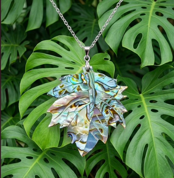 Un pendentif en forme de feuille aux reflets nacrés, irisés de bleu, vert et violet, suspendu à une fine chaîne argentée. Le bijou est mis en valeur devant un fond dense et naturel composé de grandes feuilles vertes de Monstera. L'ensemble offre un contraste harmonieux entre l'éclat métallique et nacré du pendentif et la texture végétale et colorée de la plante.