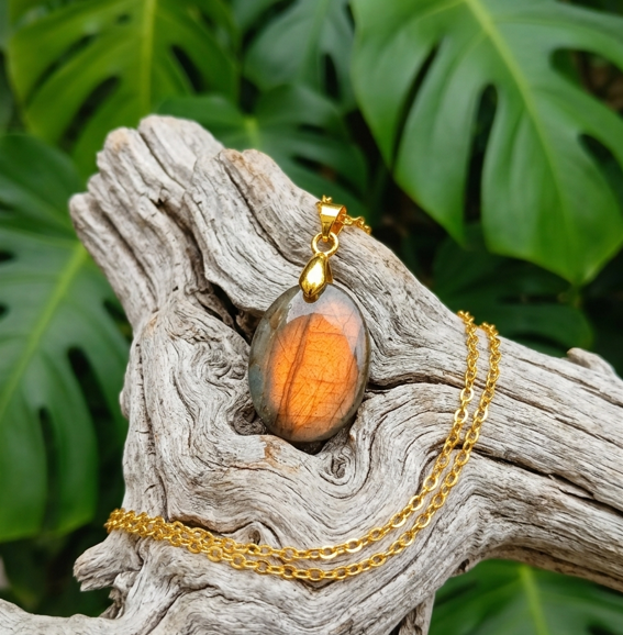 Un pendentif ovale en labradorite, dont le cœur orange scintille comme une flamme, est posé sur un morceau de bois flotté texturé. Une fine chaîne en or serpente délicatement sur le bois usé par le temps, contrastant avec la pierre lumineuse. En arrière-plan, de grandes feuilles vertes et découpées de monstera créent un décor végétal dense et apaisant.