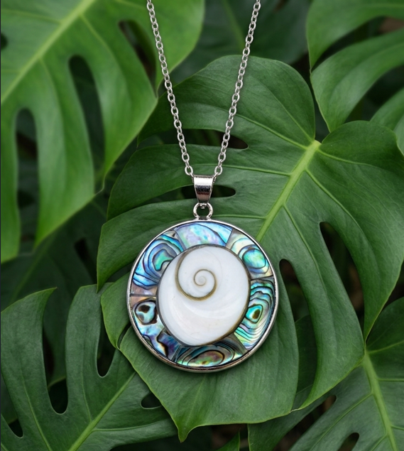 Un grand pendentif médaillon rond est présenté de face, suspendu à une fine chaîne argentée à maillons. Le bijou est délicatement posé au centre d'un arrière-plan composé de grandes feuilles vertes et fraîches de Monstera, dont les découpures naturelles entourent harmonieusement le collier. Le pendentif se compose de deux éléments naturels distincts : Au centre : Un opercule de coquillage appelé œil de Sainte-Lucie, de forme ovale et d'un blanc pur et lisse. On distingue nettement en son cœur une fine spirale brune dessinée de manière régulière. En couronne : Le coquillage central est entièrement encerclé par une large bordure en nacre abalone irisée. Cette nacre déploie des reflets métalliques intenses et changeants, mêlant des nuances de bleu roi, de turquoise, de vert émeraude et de nacre sombre. L'ensemble du médaillon est solidement enserré dans un sertissage en métal argenté brillant, surmonté d'une bélière assortie où passe la chaîne. La lumière naturelle met en valeur les contrastes entre la blancheur du centre, l'éclat coloré de la nacre et le vert végétal du fond.