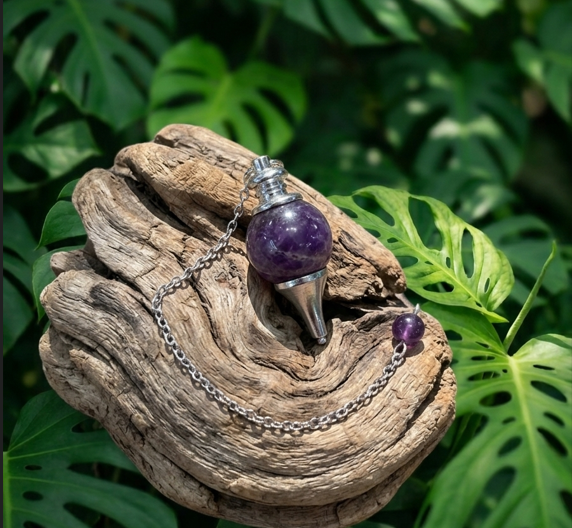 Un pendule de radiesthésie en améthyste violette, serti d'un cône en métal argenté, est posé sur un morceau de bois flotté texturé et vieilli. Une chaîne en maille argentée, également ornée d'une perle en améthyste, est reliée au pendule. Le tout est placé dans un environnement naturel et luxuriant, entouré de grandes feuilles de monstera aux découpes caractéristiques.