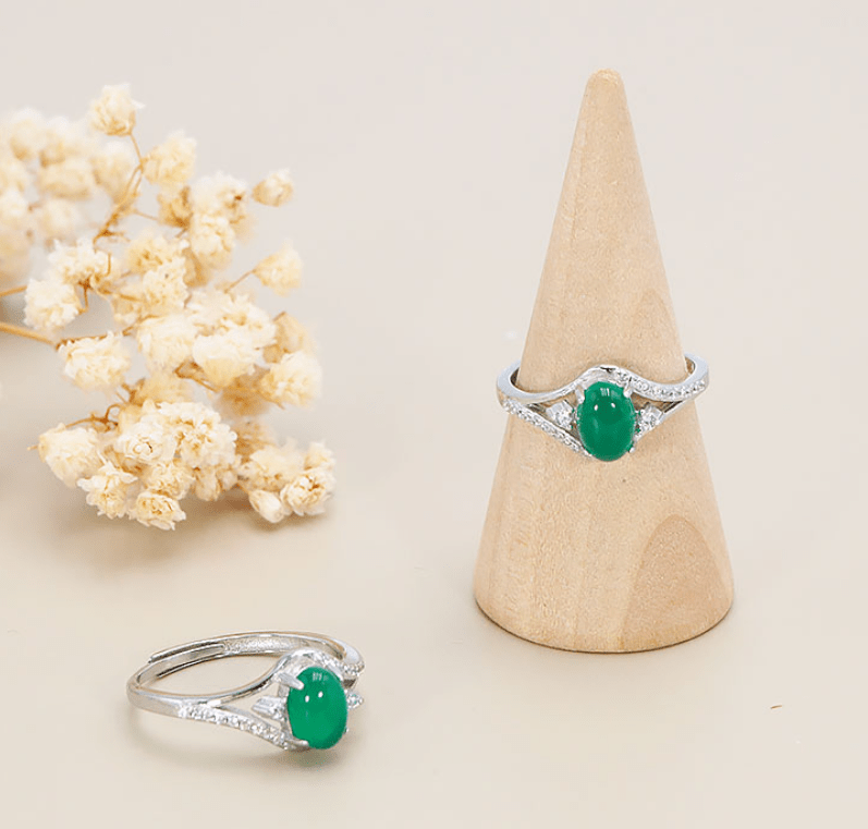 Une bague en argent ornée d'une pierre ovale vert vif, de type jade ou aventurine, polie en cabochon lisse et brillant. Serti et pierre centrale : La pierre verte est maintenue au centre par quatre griffes métalliques fines. Anneau et détails : L'anneau s'écarte en V de chaque côté du centre pour former une arche délicate. Les branches de cet anneau sont incrustées de petits strass ou brillants transparents, avec un strass légèrement plus grand posé de chaque côté de la pierre principale. L'anneau est ajustable sur le dessous. Mise en scène : Deux bagues identiques sont visibles sur un fond beige très doux. L'une est enfilée sur un cône de présentation en bois clair, l'autre est posée à plat à côté d'un petit bouquet de fleurs séchées blanches (gypsophile).