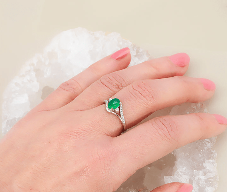 Une bague en argent ornée d'une pierre ovale vert chrysoprase portée au doigt d'une main. Serti et pierre centrale : La pierre d'un vert intense et lumineux est taillée en cabochon lisse et fixée au centre par quatre griffes métalliques. Anneau et détails : L'anneau fin s'entrouvre de chaque côté pour entourer la pierre dans un mouvement ondulé. Il est incrusté de petits strass scintillants, avec un oxyde de zirconium légèrement plus grand de chaque côté de la gemme. Mise en scène : La bague est portée à l'annuaire d'une main aux ongles vernis de rose clair. La main repose sur un bloc de quartz blanc cristallin, sur un fond beige neutre.