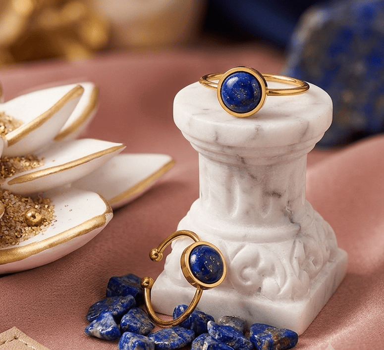 Deux bagues fines dorées serties d'une pierre ronde de lapis-lazuli d'un bleu profond sont présentées dans un décor élégant. L'une est posée au sommet d'un petit socle en marbre blanc, tandis que l'autre repose à sa base au milieu de petites pierres brutes assorties. En arrière-plan, une coupelle blanche au liseré doré et un drapé rose poudré apportent une touche chaleureuse et raffinée.