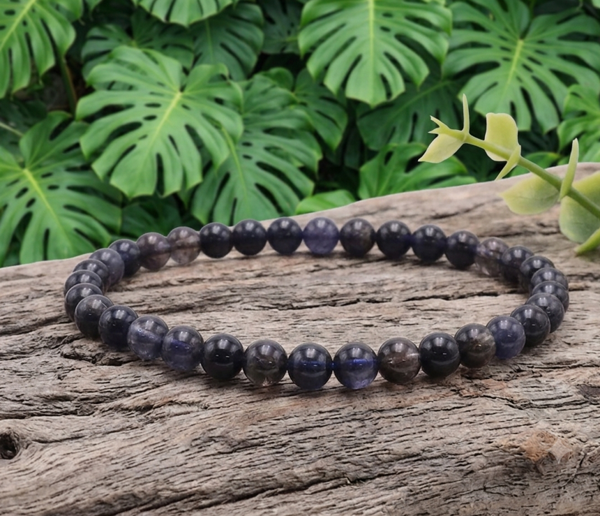 Un bracelet circulaire composé de petites perles rondes et polies de 6 mm de diamètre en cordiérite (iolite). Les perles présentent des nuances de bleu nuit, de violet translucide, de gris fumé et de noir, révélant la texture naturelle et la qualité de la pierre. Support : Le bracelet est posé à plat sur une branche de bois flotté gris clair, très texturée, dont on distingue nettement les rainures et les fibres naturelles du bois sec. Une petite tige végétale verte claire surmonte le bois sur le côté droit. Arrière-plan : Un fond végétal luxuriant composé de nombreuses feuilles vertes de Monstera, reconnaissables à leurs découpes et perforations caractéristiques, créant un décor naturel et apaisant.
