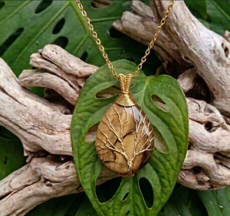 Voici une description détaillée de la dernière image générée, spécialement conçue pour les personnes malvoyantes. Cette image présente un pendentif artisanal en pierre naturelle, mis en valeur dans un décor végétal et boisé très texturé. Le pendentif La pierre : Au centre se trouve une pierre fine en forme de goutte d'eau (ou de poire inversée). Il s'agit d'une œil de tigre aux nuances chaudes et terreuses, alternant des stries horizontales et obliques marron clair, beige doré et marron foncé. La surface de la pierre est lisse et polie, reflétant doucement la lumière. Le sertissage (Arbre de vie) : La pierre est entièrement enveloppée à la main par un délicat travail de "wire wrapping" (fil de fer artistique). Un fil métallique doré de fine épaisseur part de la base pour former un tronc torsadé réaliste. De ce tronc se déploient plusieurs branches dorées qui épousent parfaitement la courbure de la pierre, créant le motif d'un arbre de vie. Les fils métalliques entourent fermement la pierre sur les côtés pour la maintenir. L'attache et la chaîne : Au sommet de la pierre, le fil doré est enroulé de manière très serrée et régulière sur plusieurs rangs pour former une bélière conique solide. Une fine chaîne dorée à maillons simples passe à travers cette attache et remonte vers le haut de l'image. Le décor de fond La feuille centrale : Le pendentif repose verticalement au centre d'une feuille verte de Monstera adansonii (souvent appelée "plante masque de singe"). Cette feuille est caractérisée par ses perforations naturelles oblongues (des trous) à travers lesquelles on devine l'arrière-plan. Elle apporte une touche de fraîcheur d'un vert moyen et présente une texture légèrement brillante. Le bois flotté : Juste derrière la feuille, on distingue des morceaux de bois flotté ou de racine ancienne, de couleur beige clair et grisâtre. Ce bois est très texturé, sec, avec des rainures et des reliefs profonds qui contrastent avec la douceur de la pierre. Le fond végétal : L'ensemble de la scène est posé sur un arrière-plan composé d'autres grandes feuilles de Monstera, créant un tapis de verdure tropicale luxuriante qui occupe tout le cadre. La lumière globale est naturelle, douce et diffuse, venant du haut pour mettre en valeur les reflets dorés du fil métallique et les détails de la pierre.