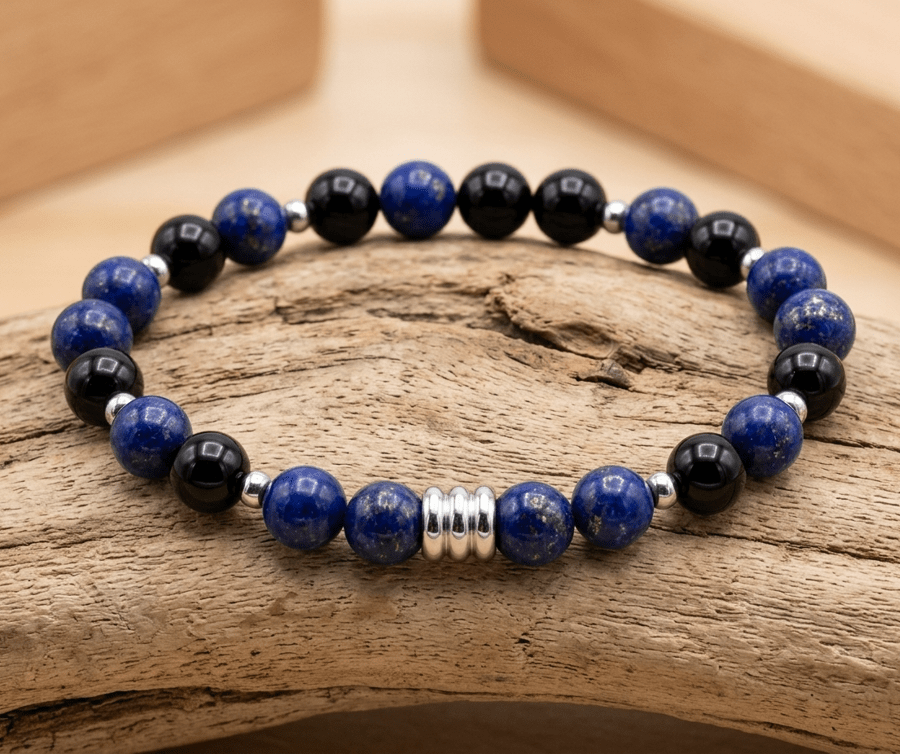 Un bracelet en perles rondes en pierre naturelle posé à plat sur une branche de bois flotté texturé, dans un décor épuré aux tons de bois clair d'inspiration Montessori. Composition du bracelet : Il alterne des perles de lapis lazuli d'un bleu nuit profond parsemé de minuscules paillettes dorées (pyrite) et des perles d'onyx noir parfaitement lisses et brillantes. Détails métalliques : De très fins intercalaires en métal argenté séparent chaque groupe de perles. Au centre du bracelet trône un élément tubulaire argenté brillant strié de trois anneaux en relief. Arrière-plan : Le bois flotté repose sur un plateau en bois blond baigné d'une lumière douce et naturelle, créant une atmosphère sereine et chaleureuse.