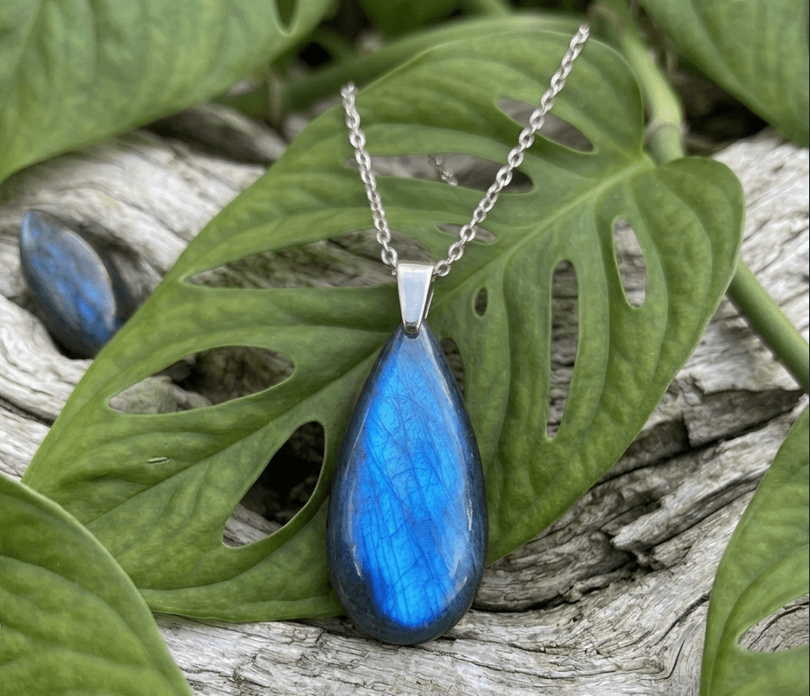 Cette image présente un pendentif en forme de goutte, fabriqué à partir d'une pierre de labradorite bleue polie. La pierre capte la lumière et affiche des couleurs bleues vibrantes et iridescentes. Le pendentif est fixé à une fine chaîne argentée et repose sur une surface en bois texturé et vieilli. En arrière-plan, plusieurs feuilles de monstera vertes ajourées sont visibles, encadrant le pendentif et ajoutant une touche de nature à l'image.
