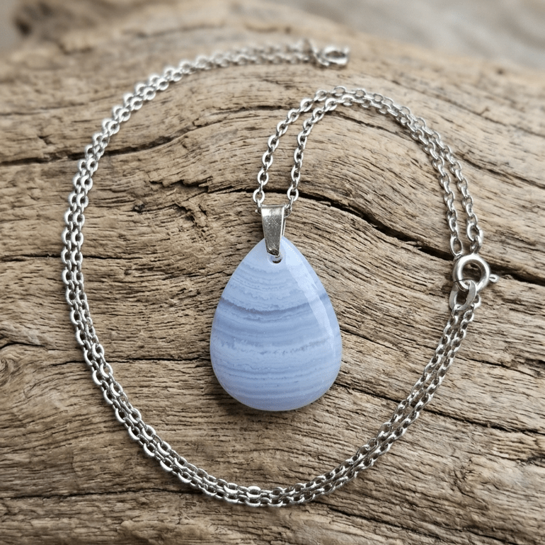Pendentif pierre calcédoine en vue rapprochée d'un collier délicat en calcédoine bleue de Namibie, reposant sur un morceau de bois flotté texturé. La chaîne en acier inoxydable à mailles s'enroule gracieusement autour du pendentif en forme de goutte, qui présente de subtiles bandes horizontales bleu clair et lilas. Le pendentif est suspendu par une petite bélière argentée et simple. Le fermoir à ressort se trouve sur le côté droit de la chaîne. L'arrière-plan en bois flotté présente des textures boisées riches et vieillies, ajoutant de la profondeur à l'image.