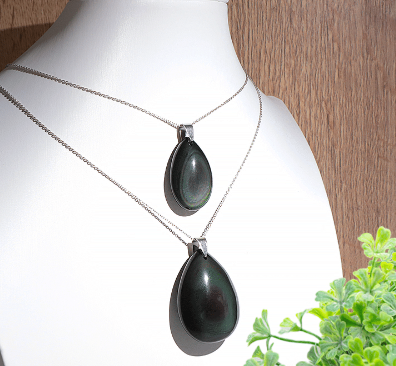deux pendentifs en obsidienne œil céleste en forme de goutte d'eau lisse et polie, présentés l'un au-dessus de l'autre sur un buste d'exposition blanc. Le pendentif du haut, plus petit (35 mm), et celui du bas, plus grand (50 mm), sont suspendus à de fines chaînes argentées. La pierre noire polished révèle de subtils reflets irisés et concentriques aux nuances vertes et violettes sous la lumière. L'ensemble est mis en valeur par un fond en bois clair et quelques petites feuilles vertes disposées au premier plan à droite.