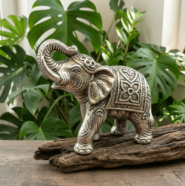 Une photo carrée présente une petite statuette de 6 cm représentant un éléphant en métal argenté vieilli. L'éléphant est représenté de profil, la trompe levée vers le haut en signe de chance, drapé d'un tapis de selle richement gravé de motifs floraux et traditionnels. La sculpture repose sur une pièce de bois flotté usé, sur un fond apaisant de grandes feuilles de Monstera éclairées d'une lumière naturelle douce.