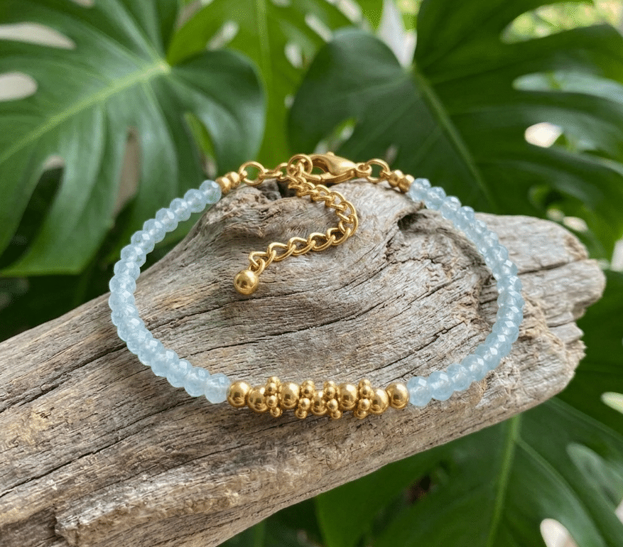 bracelet délicat posé sur un morceau de bois flotté gris et texturé. Le bracelet est composé de petites perles d'aigue-marine bleu clair facettées et de plusieurs perles dorées lisses regroupées au centre. Une chaîne d'extension dorée avec un fermoir mousqueton est visible à l'arrière. L'arrière-plan est constitué de grandes feuilles vertes de Monstera floues