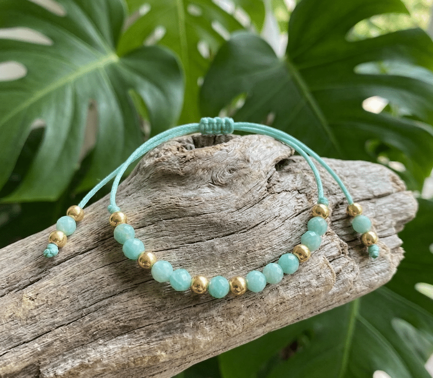 Un bracelet en cordon vert d'eau est délicatement posé sur un morceau de bois flotté texturé et patiné. Le bracelet est orné d'une rangée de perles à facettes en pierre d'Amazonite d'un vert bleuté doux, alternées avec de petites billes métalliques dorées polies. En arrière-plan, hors focus, de grandes feuilles vertes de Monstera aux découpes caractéristiques créent une ambiance naturelle et tropicale. Le cordon du bracelet est tressé et se termine par des nœuds coulissants ornés de petites perles d'Amazonite et dorées aux extrémités. L'éclairage est naturel et doux.