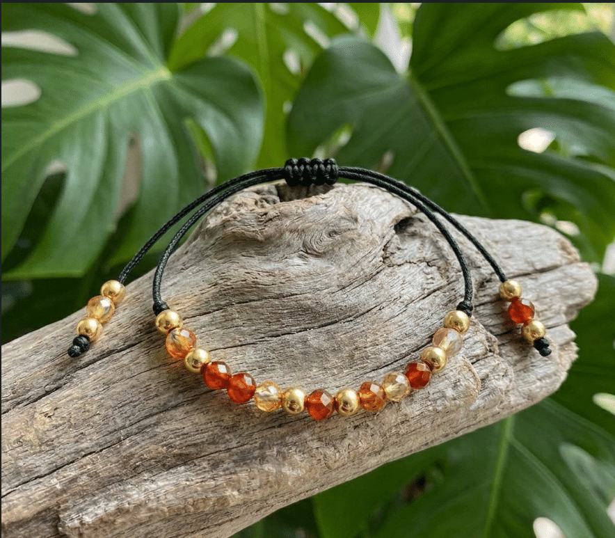 Bracelet de lithothérapie en pierre naturelle de Cornaline aux nuances orange et rouge feu, monté sur un cordon noir réglable avec des perles dorées. Le bijou est élégamment présenté sur du bois flotté naturel, sur un fond de grandes feuilles vertes de Monstera au style tropical et végétal.