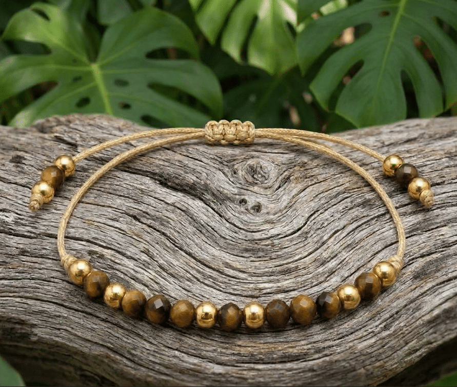 Bracelet de lithothérapie en pierre naturelle d'Œil de Tigre aux teintes marron et dorées, monté sur un cordon beige résistant avec nœud macramé coulissant. Le bracelet alterne des perles de pierre facettées et des perles en acier inoxydable dorées. Il est disposé sur un morceau de bois flotté texturé avec un arrière-plan végétal composé de feuillage de Monstera.