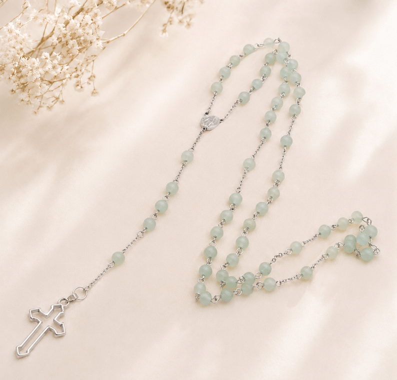 chapelet étalé avec élégance sur une pièce de bois flotté texturée. Le chapelet est composé de perles sphériques polies en aventurine verte d'un vert d'eau délicat et translucide, reliées par des maillons fins en acier inoxydable argenté. Il se termine par une médaille centrale et une croix latine en métal argenté. En arrière-plan, de grandes feuilles vertes de Monstera apportent un cadre naturel apaisant sous une lumière douce.
