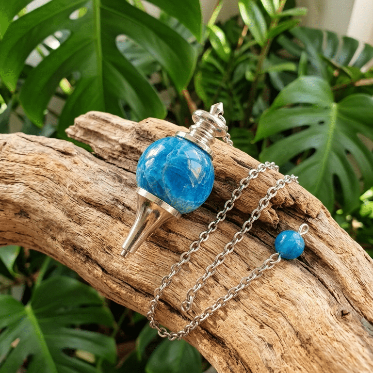 Une photo carrée montre, en gros plan et en plongée, un pendule de radiesthésie en apatite bleue. La pierre, une sphère polie bleu vif mouchetée, est sertie dans une monture métallique argentée en forme de cône inversé. Elle est posée sur un morceau de bois flotté usé et texturé. Une fine chaîne en acier s'étire depuis la monture en formant une boucle lâche vers la droite, se terminant par une petite perle d'apatite assortie. L'arrière-plan est un fouillis flou de grandes feuilles vertes de Monstera et d'autres plantes tropicales, baignées d'une lumière naturelle douce.