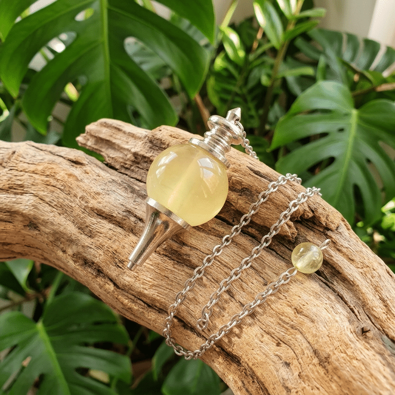 Une photo carrée présente un pendule en citrine naturelle jaune clair translucide, constitué d'une sphère polie montée sur une structure métallique argentée. Le pendule repose sur un morceau de bois flotté usé aux tons chauds. Sa chaînette en métal argenté s'étire doucement avec une petite perle de citrine à son extrémité. En arrière-plan, un feuillage vert de Monstera apporte une touche naturelle et apaisante sous une lumière douce.